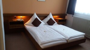 A double bed with white linens and two brown pillows, a bedside table with a glass of red wine, and a nightstand with a lamp.