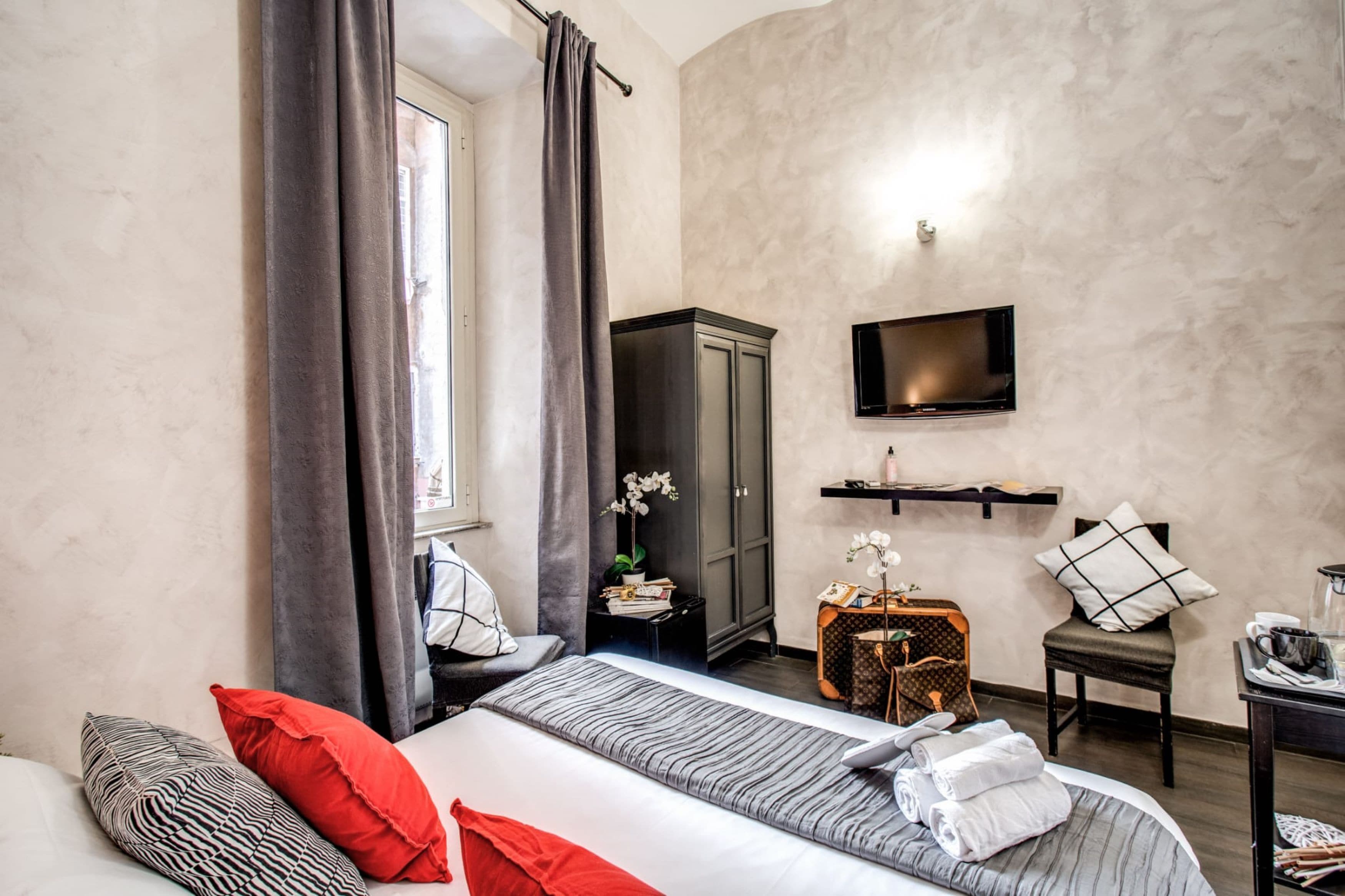 A modern hotel room with a bed, a TV mounted on the wall, a wardrobe, and a small table with a lamp.