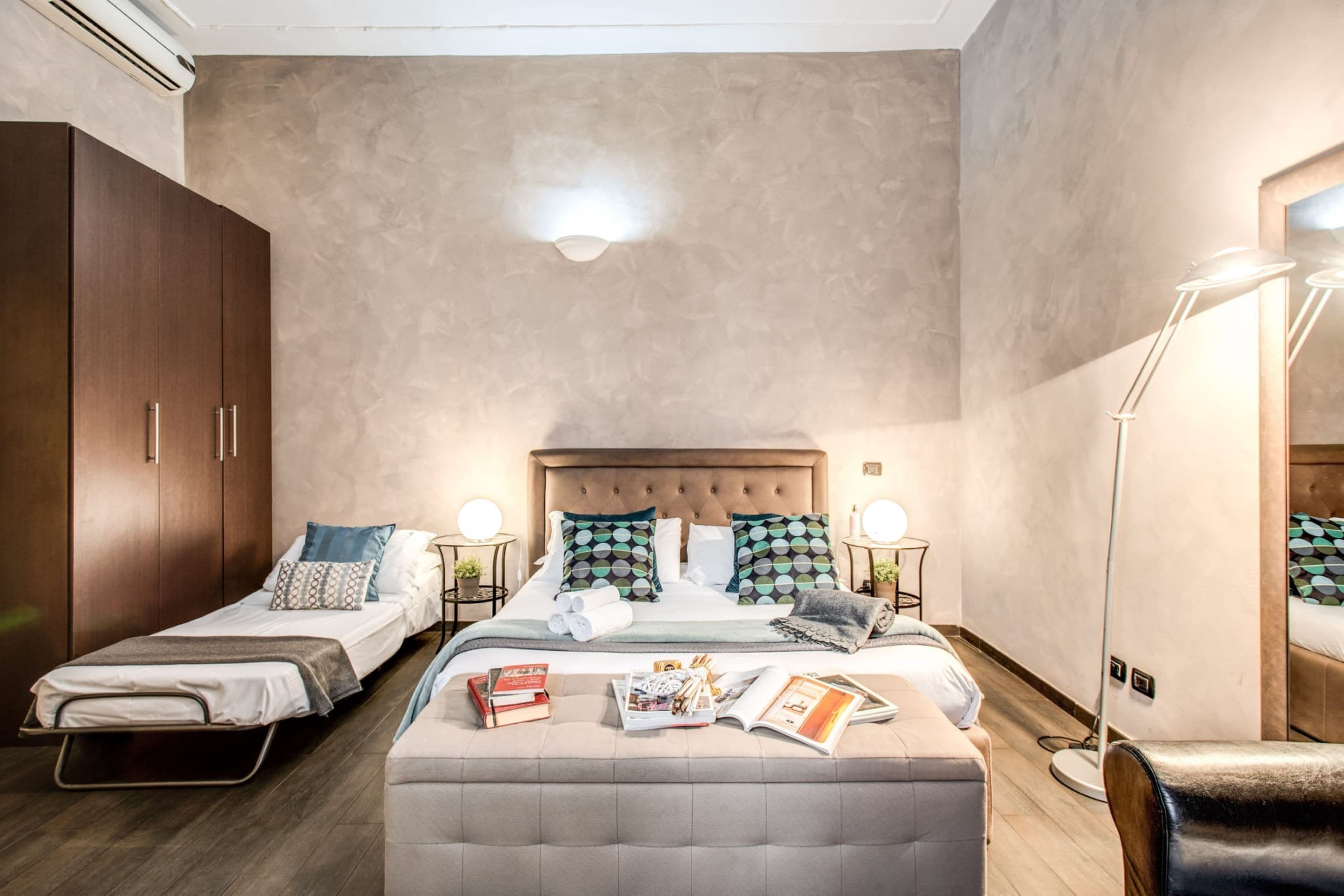 A bedroom with a bed, two bedside tables, a wardrobe, a floor lamp, and a reading area with books and magazines.