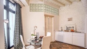 A dining area with a glass table, white chairs, a vase with flowers, and a kitchen with white cabinets and a tiled backsplash. - La Residenza dell'Angelo (Rome)