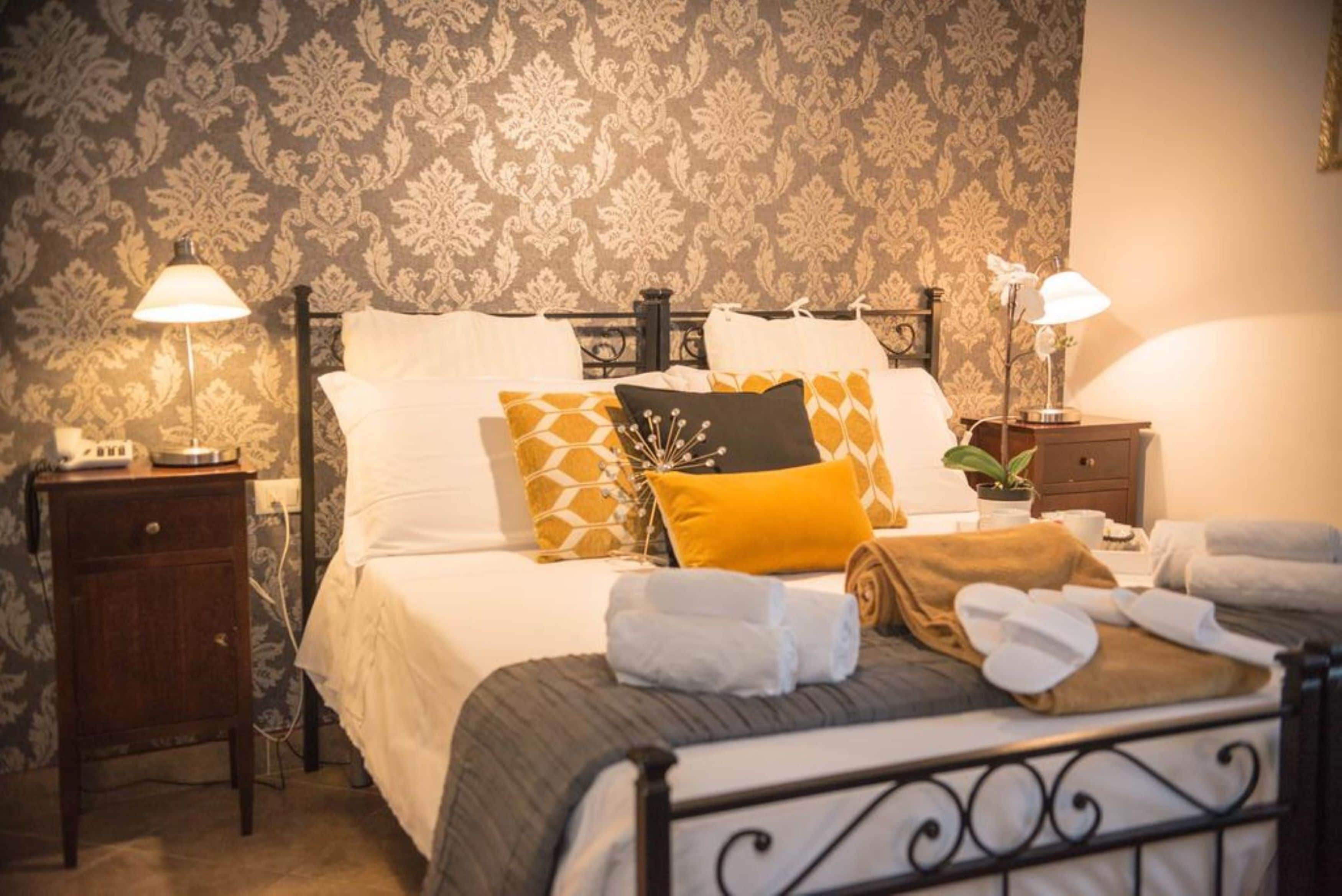 Una cama bien hecha con almohadas blancas y estampadas, una lámpara de noche y una mesita de noche con un teléfono y una planta.