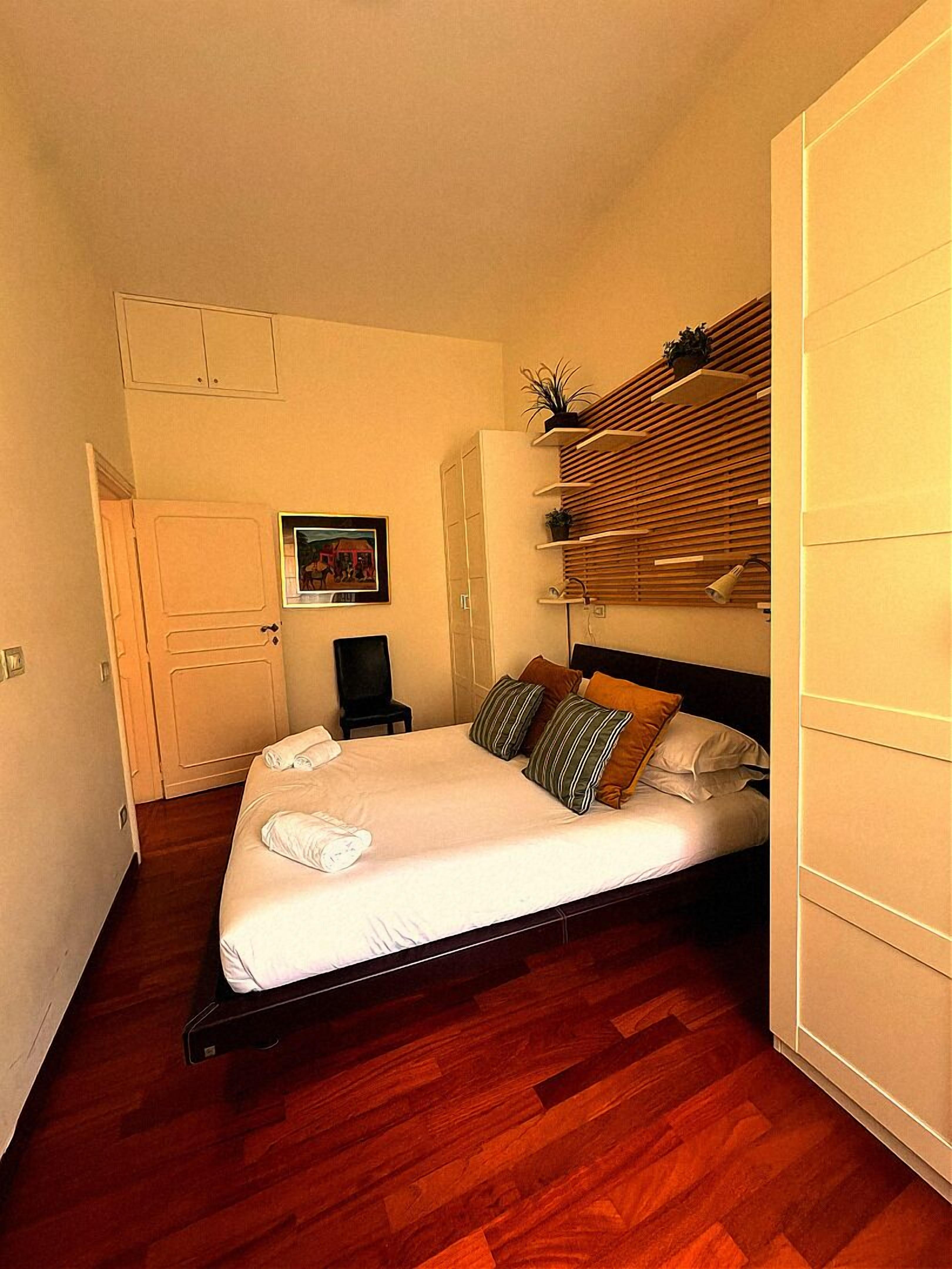 A bedroom with a bed, wooden floor, a chair, a desk, a wardrobe, and a picture on the wall.