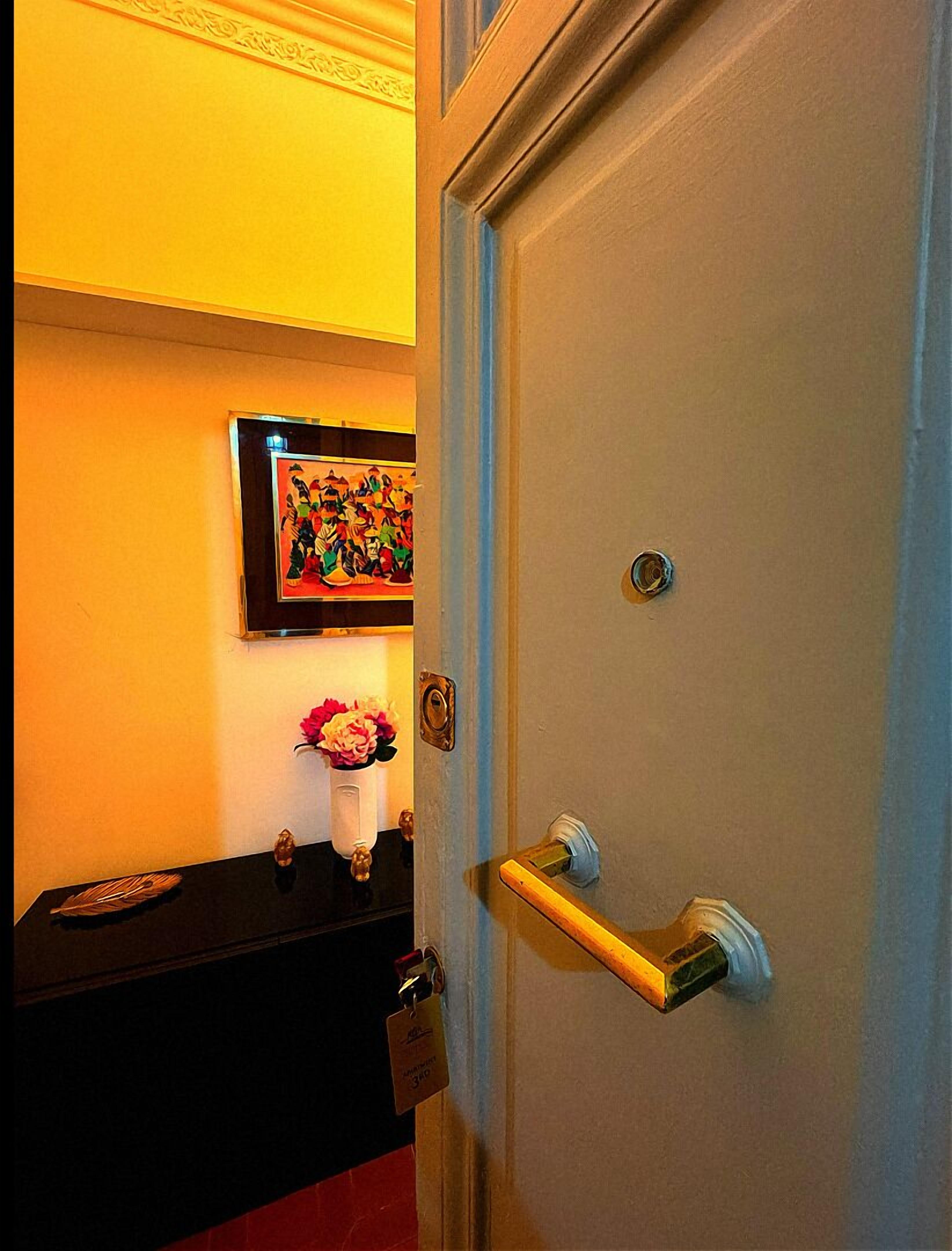 A hotel room with a door, a framed artwork on the wall, a small table with a vase of flowers, and a keychain on the door handle.