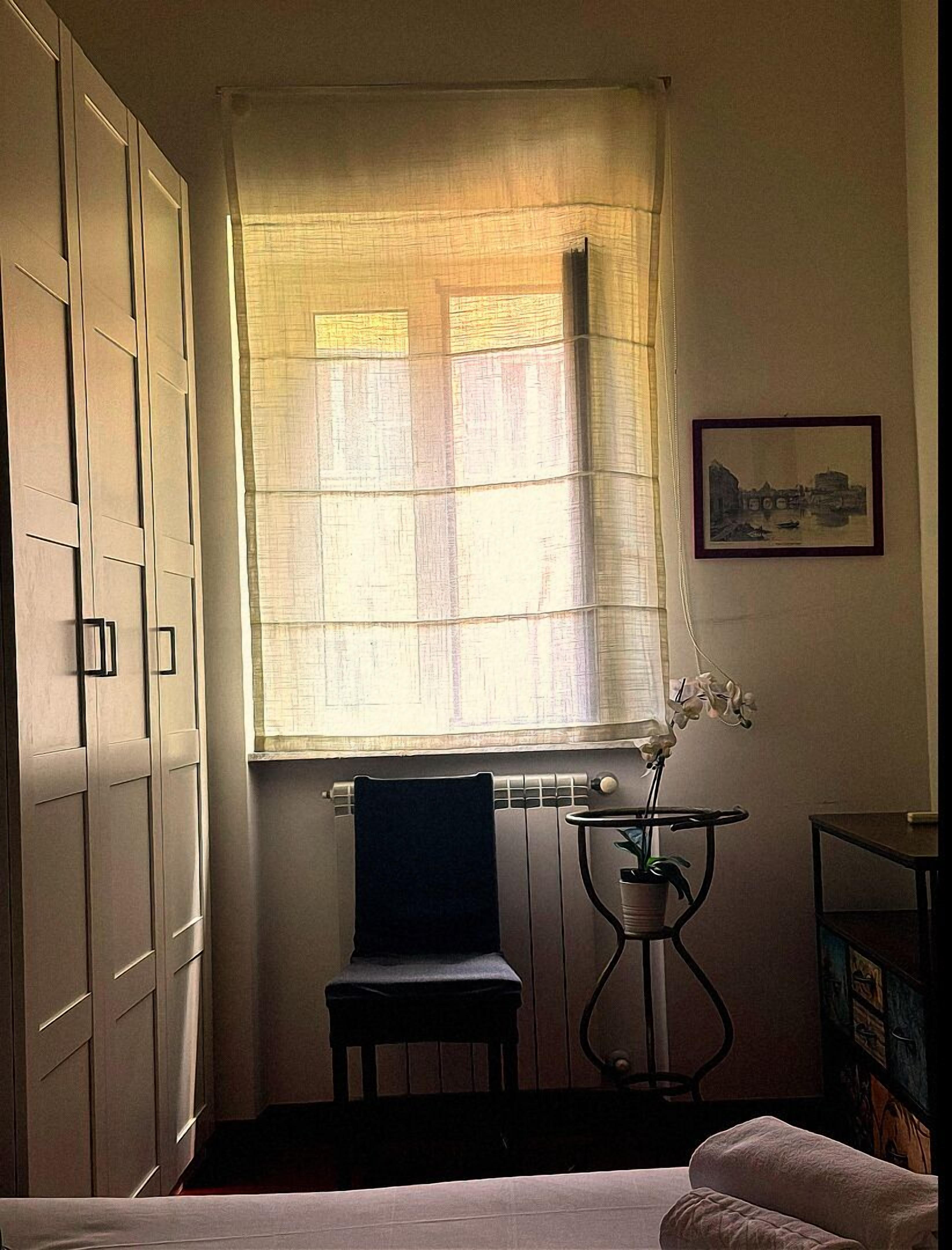 A room with a bed, a chair, a radiator, a window with sheer curtains, and a framed picture on the wall.