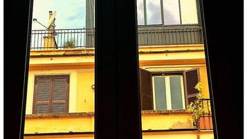 Deux fenêtres avec volets, un balcon orné de plantes et une façade d’immeuble aux couleurs jaune et orange.