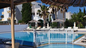 A swimming pool with lounge chairs and umbrellas, surrounded by palm trees and buildings. - Zouboulia (Kos)