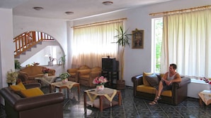 A spacious living room with a high ceiling, a staircase, and a large window with curtains. - Pagona Hotel (Istiaia-Aidipsos)