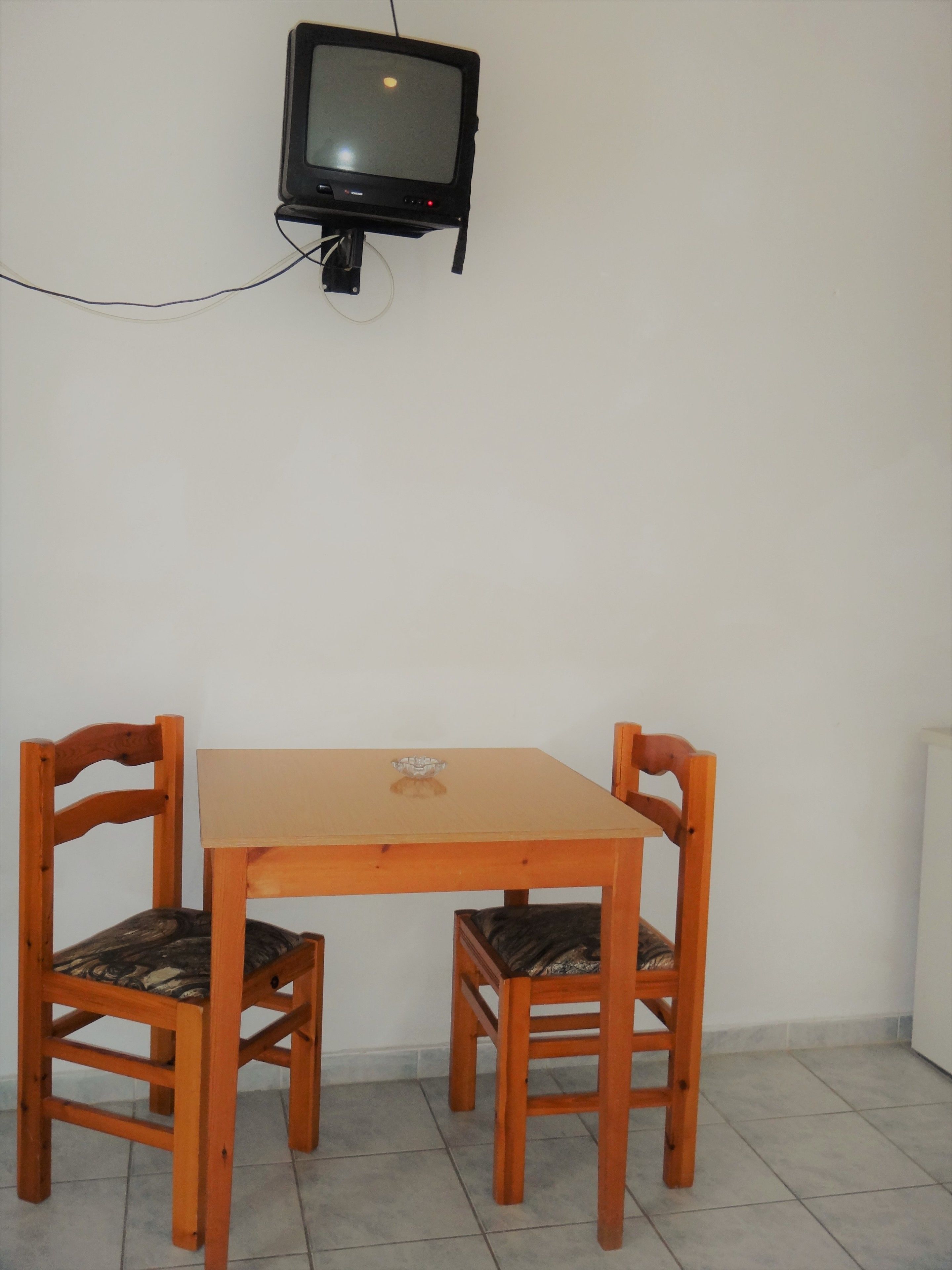 Una habitación pequeña con una mesa de madera y dos sillas, un televisor montado en la pared y un radiador en el suelo.