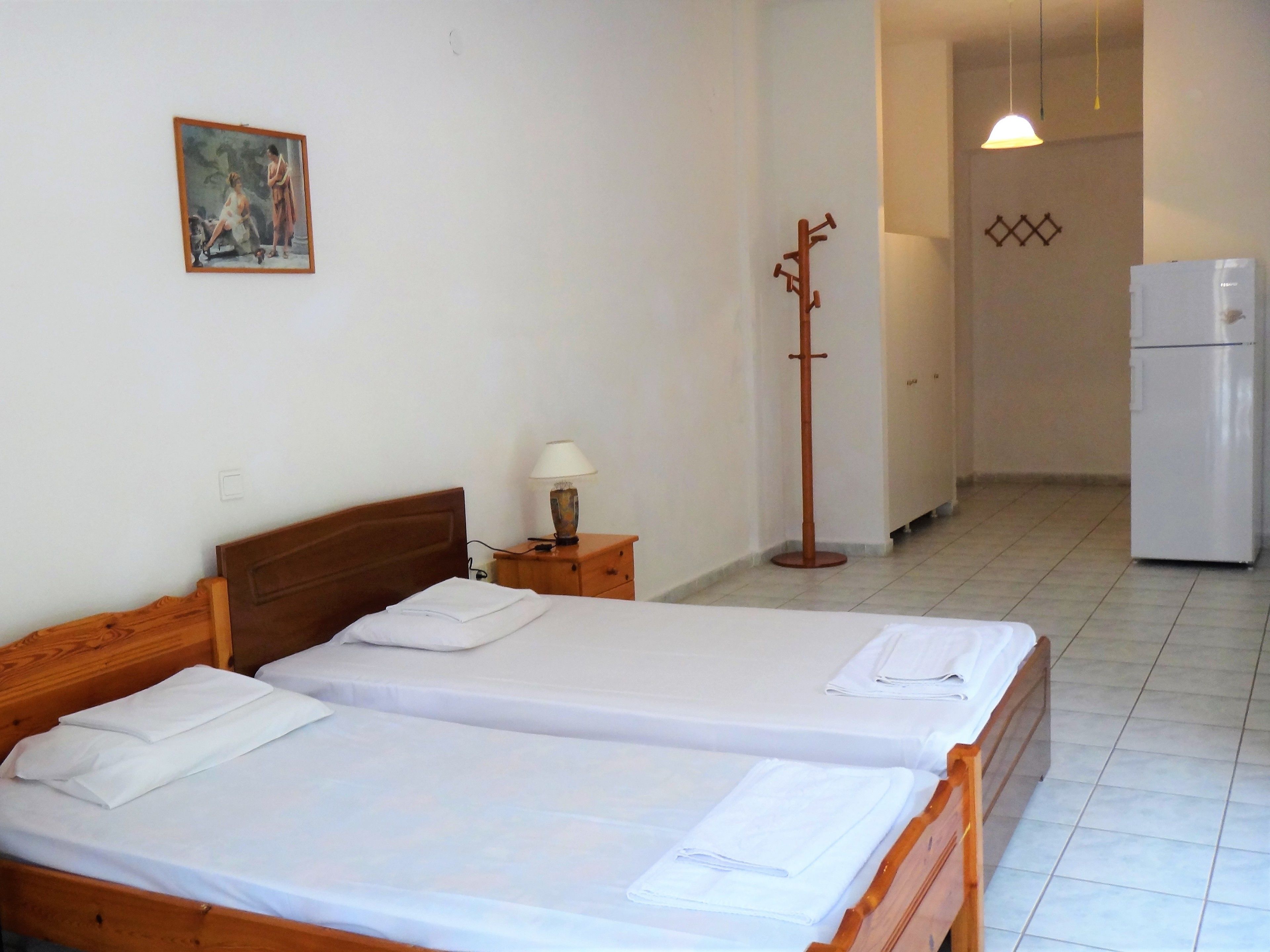 A hotel room with two wooden beds, a nightstand with a lamp, a coat rack, a refrigerator, and a painting on the wall.
