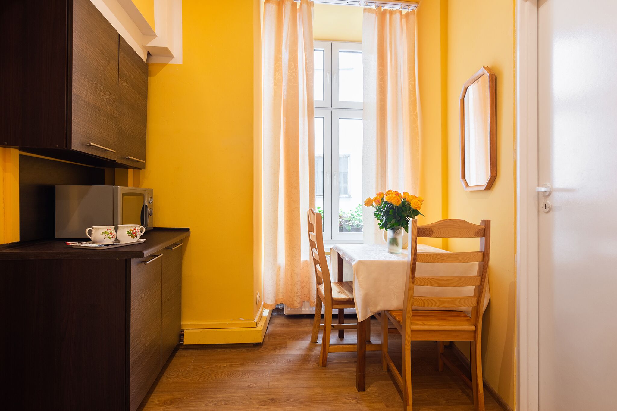 Una cucina compatta con forno a microonde, un piccolo tavolo con un vaso di fiori e una sedia di legno.