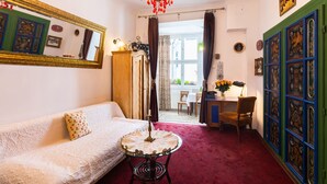 A room with a white sofa, a round table with a lace tablecloth, a chandelier, and a view of the outside through a window with curtains. - Unikat Guest Rooms (Self-check in) (Kraków)