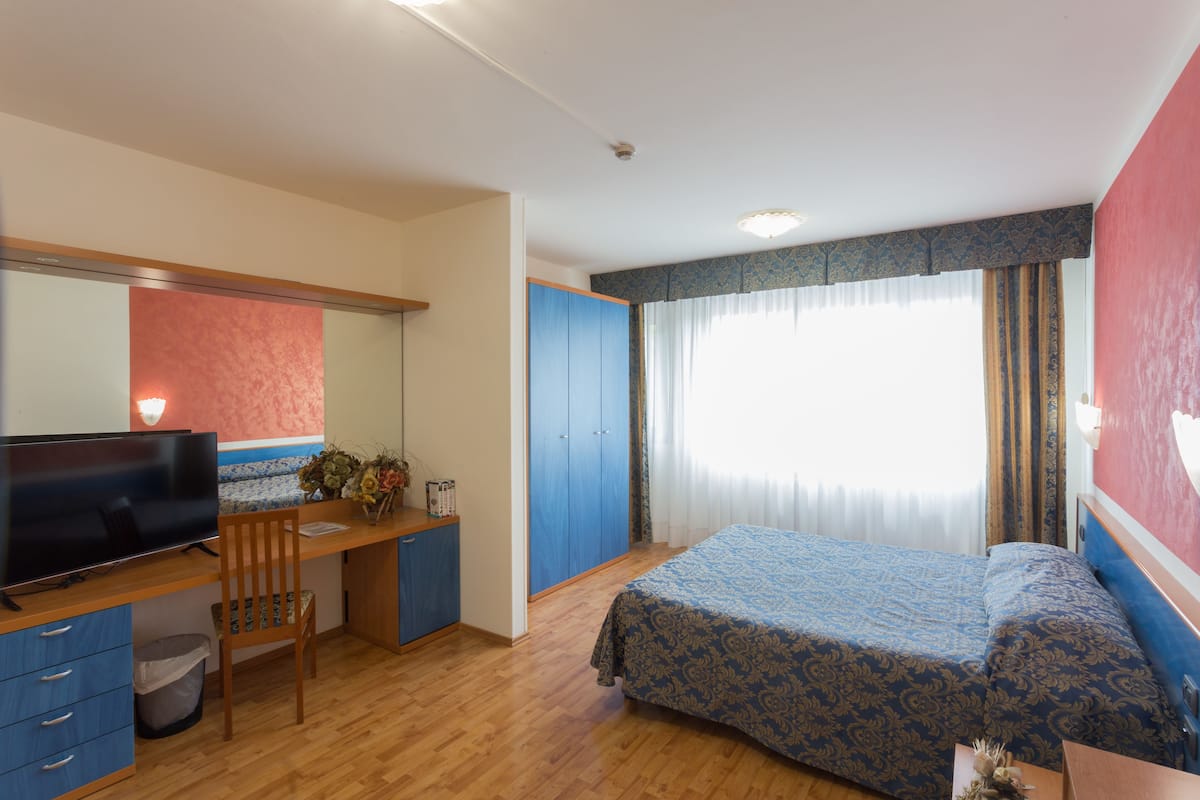 Un dormitorio con cama, un escritorio con computadora, una silla, un televisor y una ventana con cortinas.