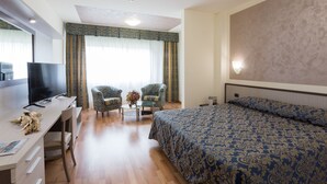 A hotel room with a bed, a desk with a TV, two armchairs, a small table with a vase, and a large window with curtains. - Hotel Airmotel (Mestre)