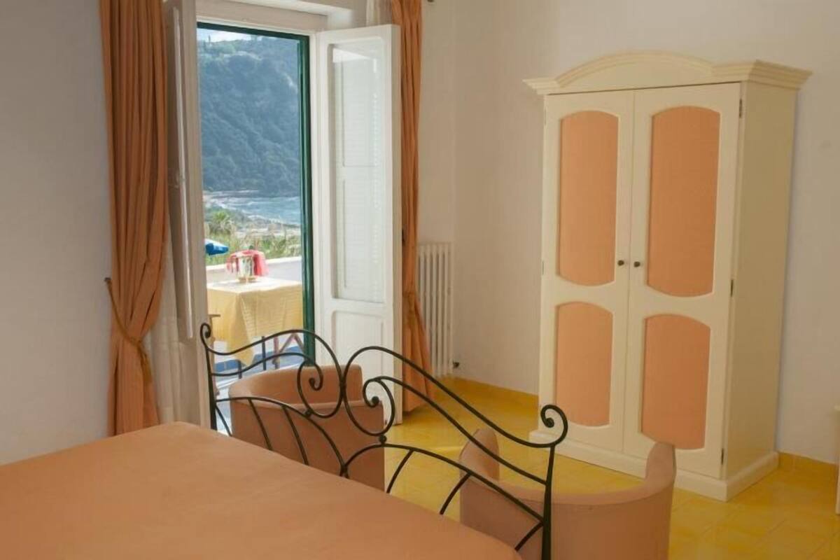 Ein Schlafzimmer mit Bett, Kleiderschrank und Blick auf eine gebirgige Landschaft durch eine offene Tür.