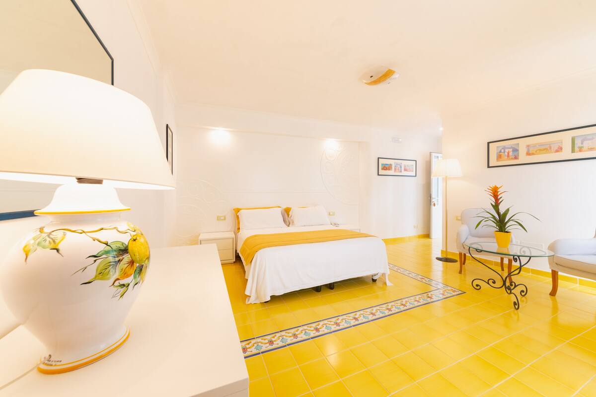Ein Hotelzimmer mit einem Bett, einem Nachttisch mit Lampe, einem kleinen Tisch mit Pflanze und einem gelb gefliesten Boden.