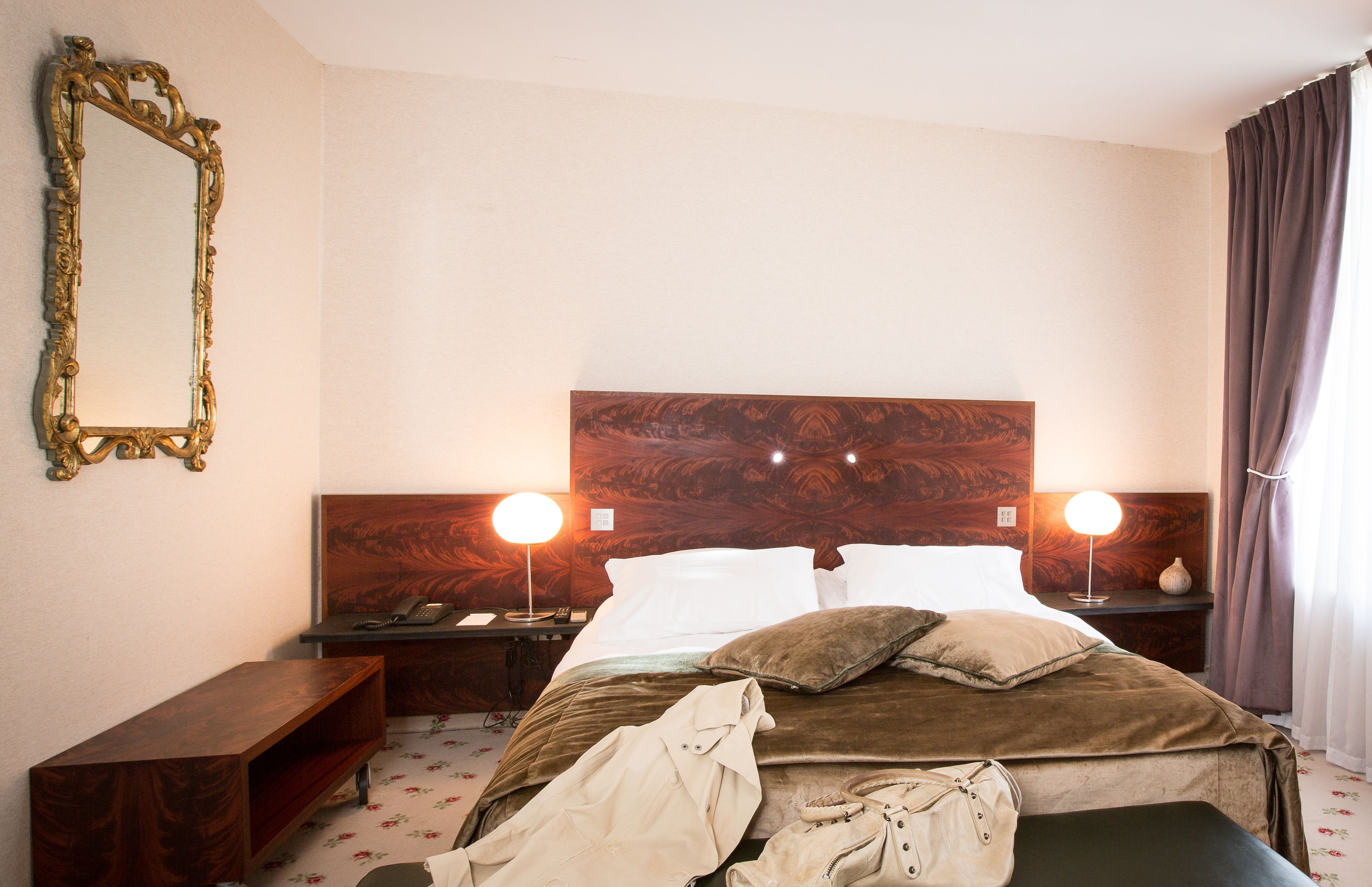 A hotel room with a wooden headboard, two bedside lamps, a nightstand, a mirror, a bed with pillows, and a bedside table.