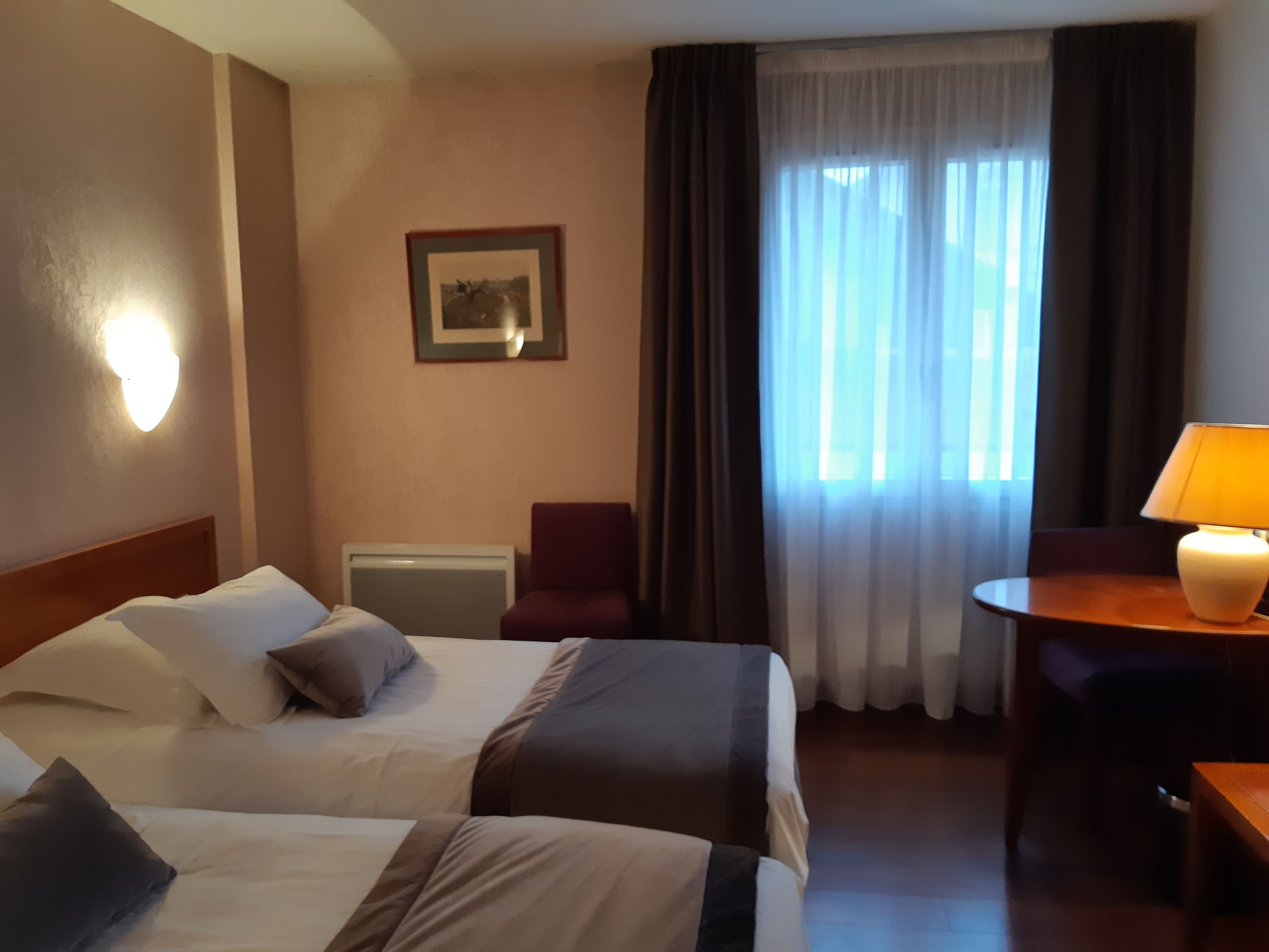 Een hotelkamer met twee bedden, een bureau, een stoel, een lamp, een schilderij aan de muur en een raam met gordijnen.