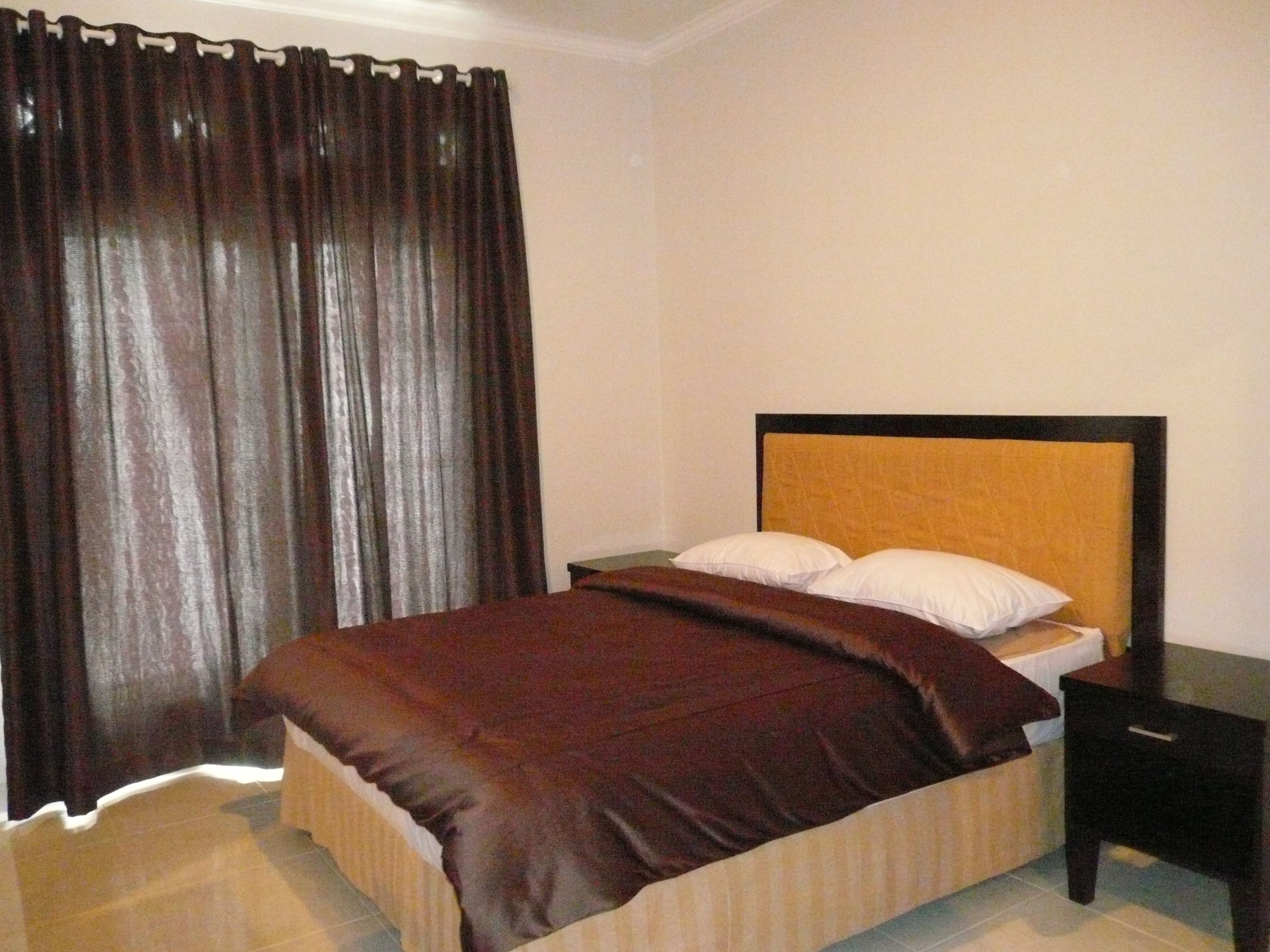 Una cama bien hecha con un edredón oscuro y almohadas, un cabecero de madera y una mesita de noche con una lámpara.
