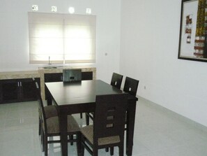 A dining area with a rectangular table and six chairs, a window with blinds, and a framed picture on the wall. - The Radiant Villa's & Function Hall (Lembang)