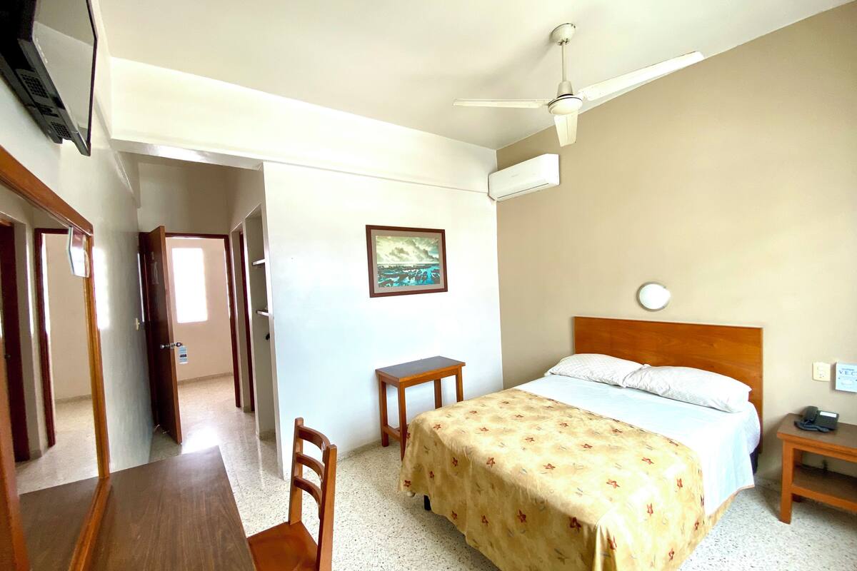 A bedroom with a bed, a desk, a chair, a ceiling fan, and a painting on the wall.