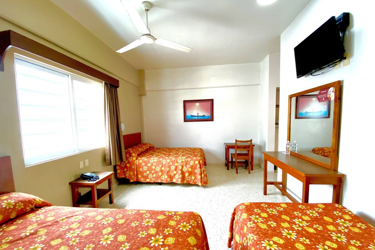 A hotel room with two beds, a desk, a chair, a ceiling fan, a TV, and a window with curtains.