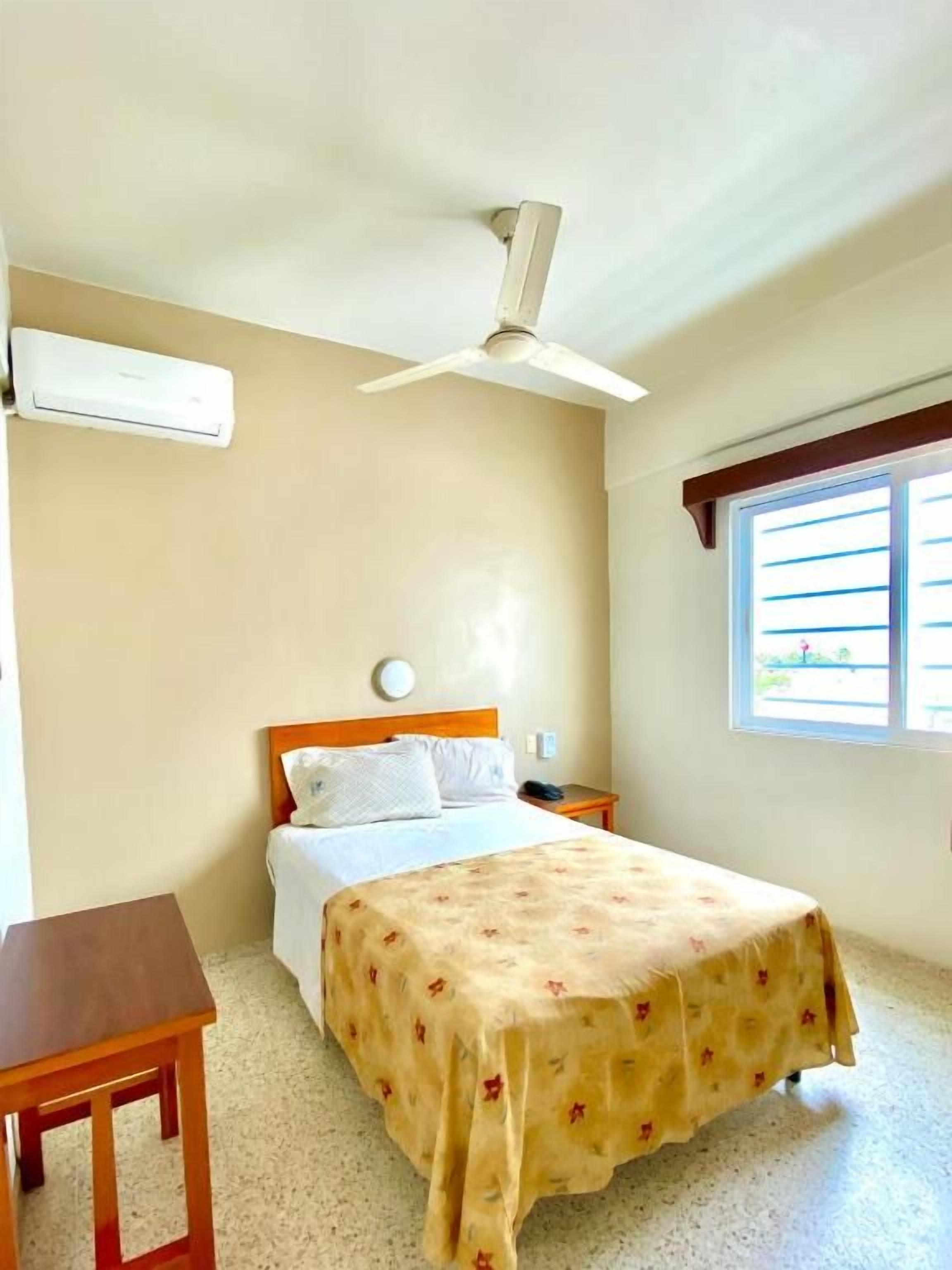 Een hotelkamer met een bed, nachtkastje, jaloezieën, plafondventilator en een airconditioning.