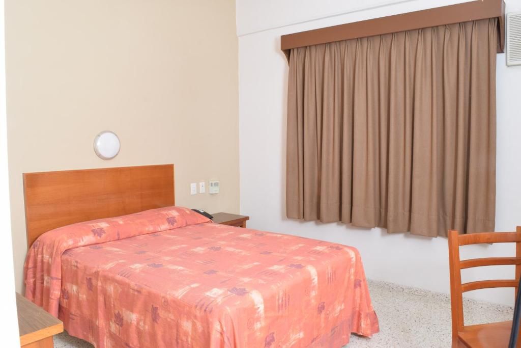 Una camera d'albergo con un letto, una testiera in legno, un comodino, una sedia e una finestra con tende.