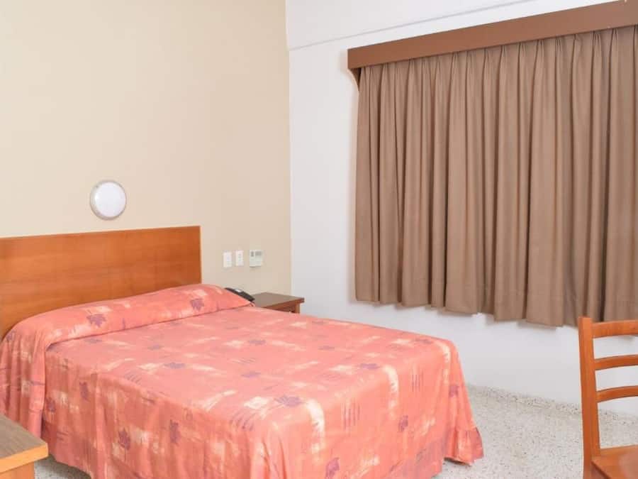 Habitación de hotel con cama, cabecera de madera, mesita de noche, una silla y ventana con cortinas.