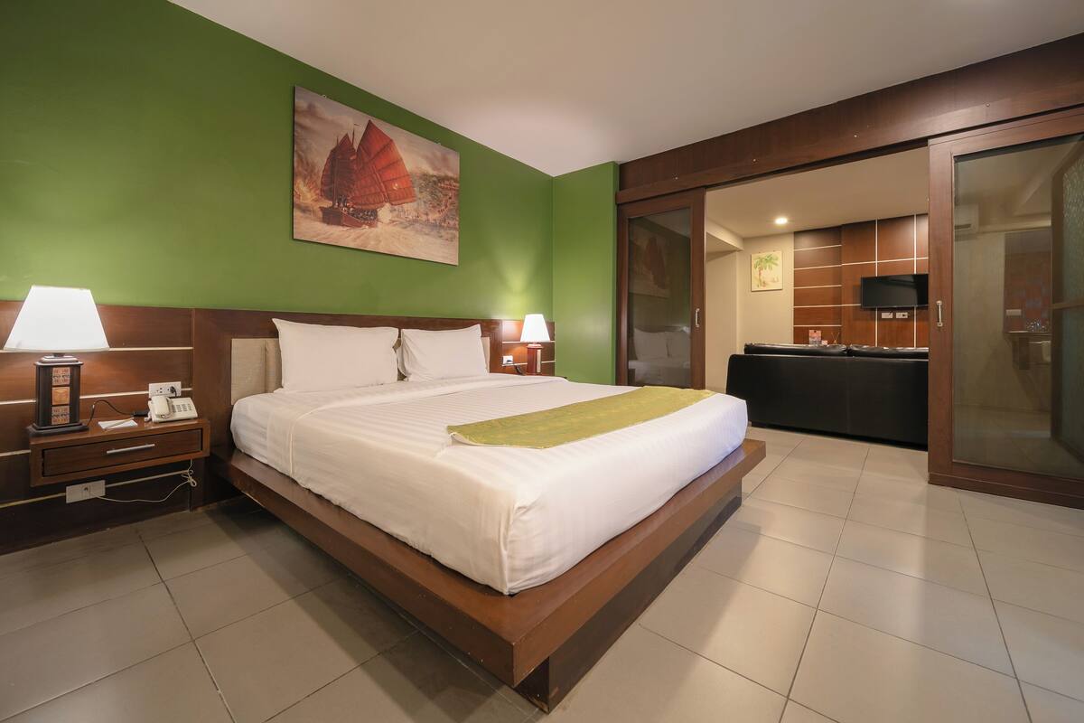 A hotel room with a large bed, two bedside lamps, a green wall, a painting, a sliding door to a kitchenette, and a TV.