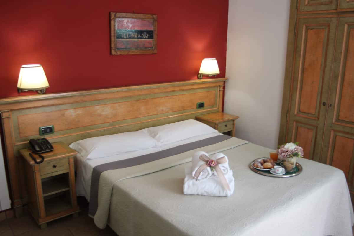 Ein Hotelzimmer mit einem hölzernen Kopfteil, zwei Nachttischlampen, einem Nachttischtelefon, einem ordentlich bezogenen Bett mit weißer Decke und einem Tablett mit Frühstücksartikeln. An der Wand hängt ein Gemälde.