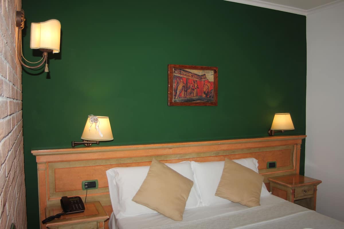 Ein Hotelzimmer mit grüner Wand, einem Holzkopfteil, einem Bett mit weißen Laken und beigen Kissen, zwei Nachttischlampen, einem Nachttisch mit Telefon und einem gerahmten Bild über dem Bett.