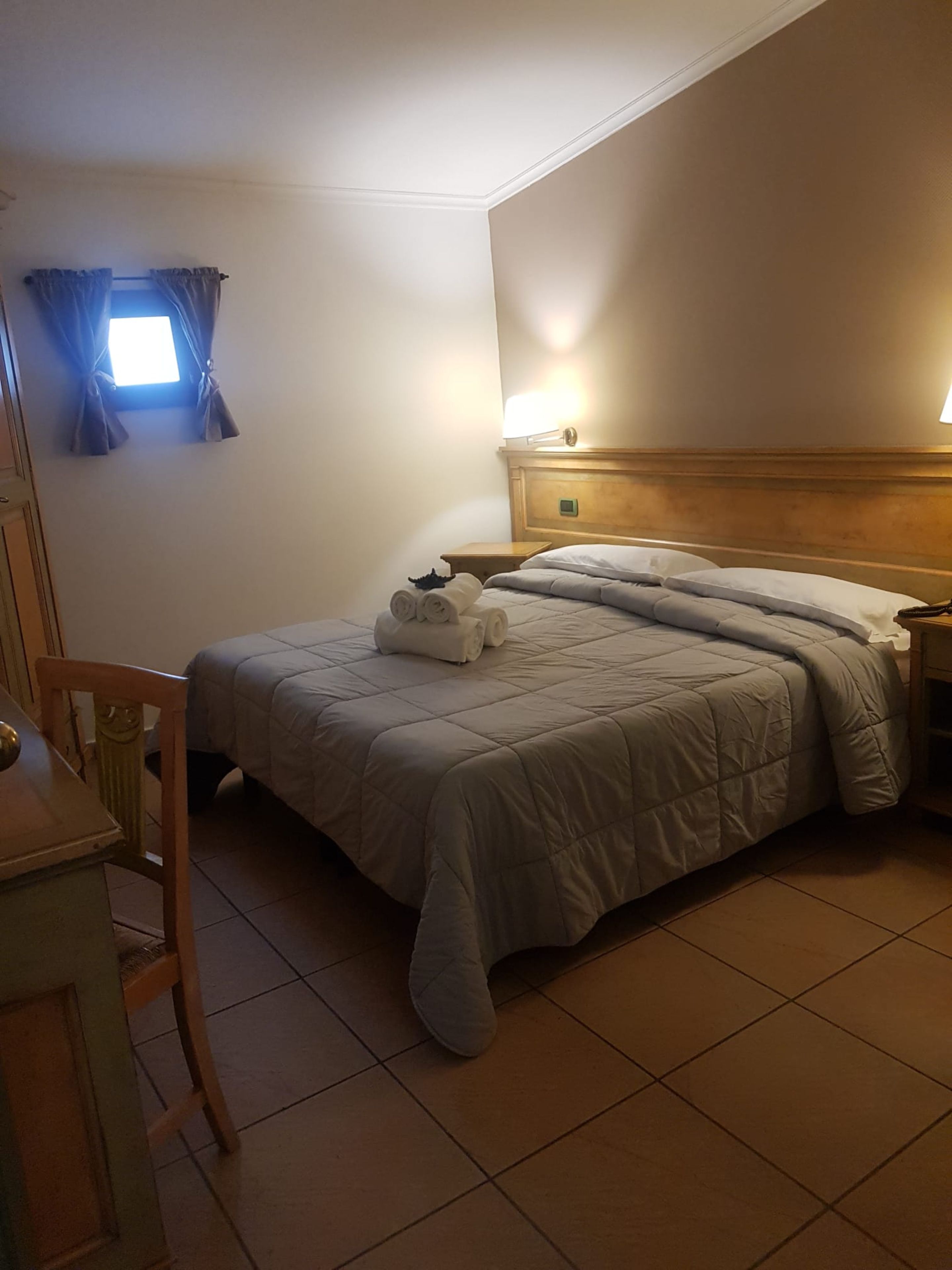 A hotel room with a bed, a chair, a small table, and a window with curtains.
