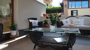 Mesa com tampo de vidro, cadeiras de vime, um sofá com almofadas e plantas em vasos num terraço.