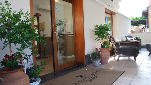 Entrada de hotel com tapete, plantas em vasos, mobiliário de vime e porta de vidro.