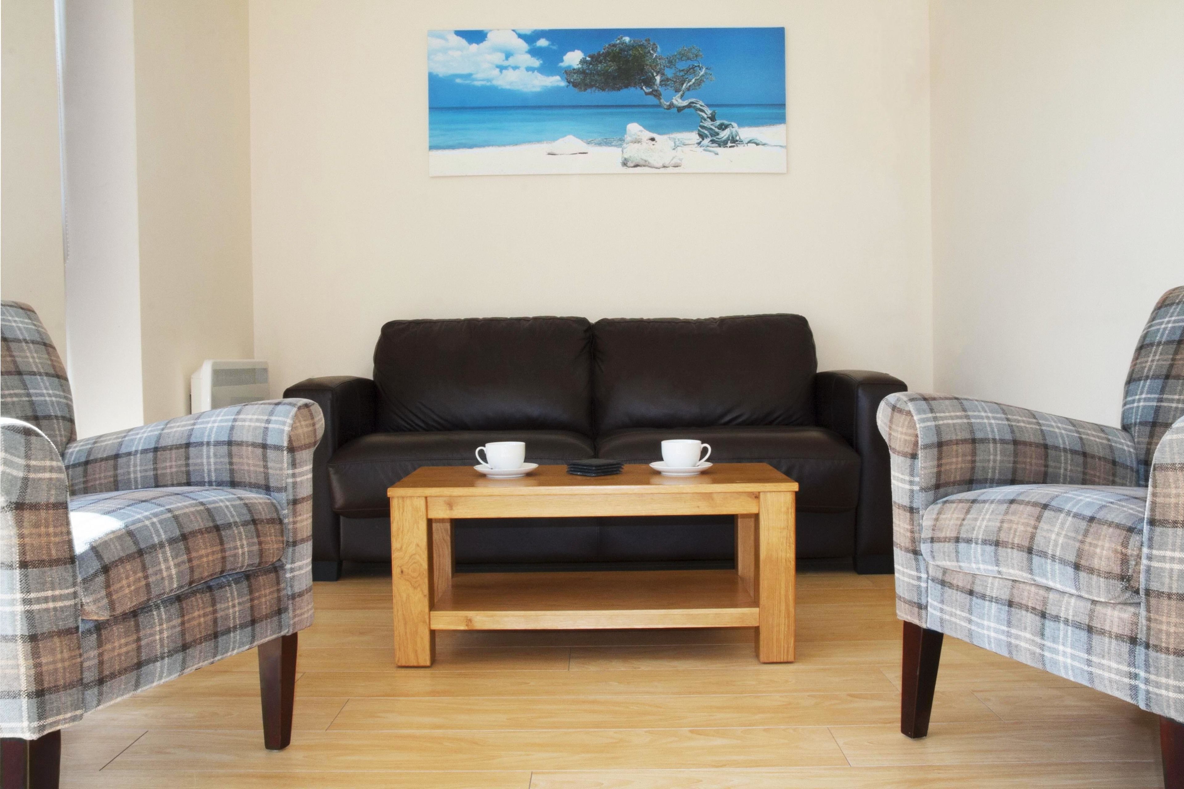 A living room with a dark brown sofa, two plaid armchairs, a wooden coffee table, and a painting of a beach scene on the wall.
