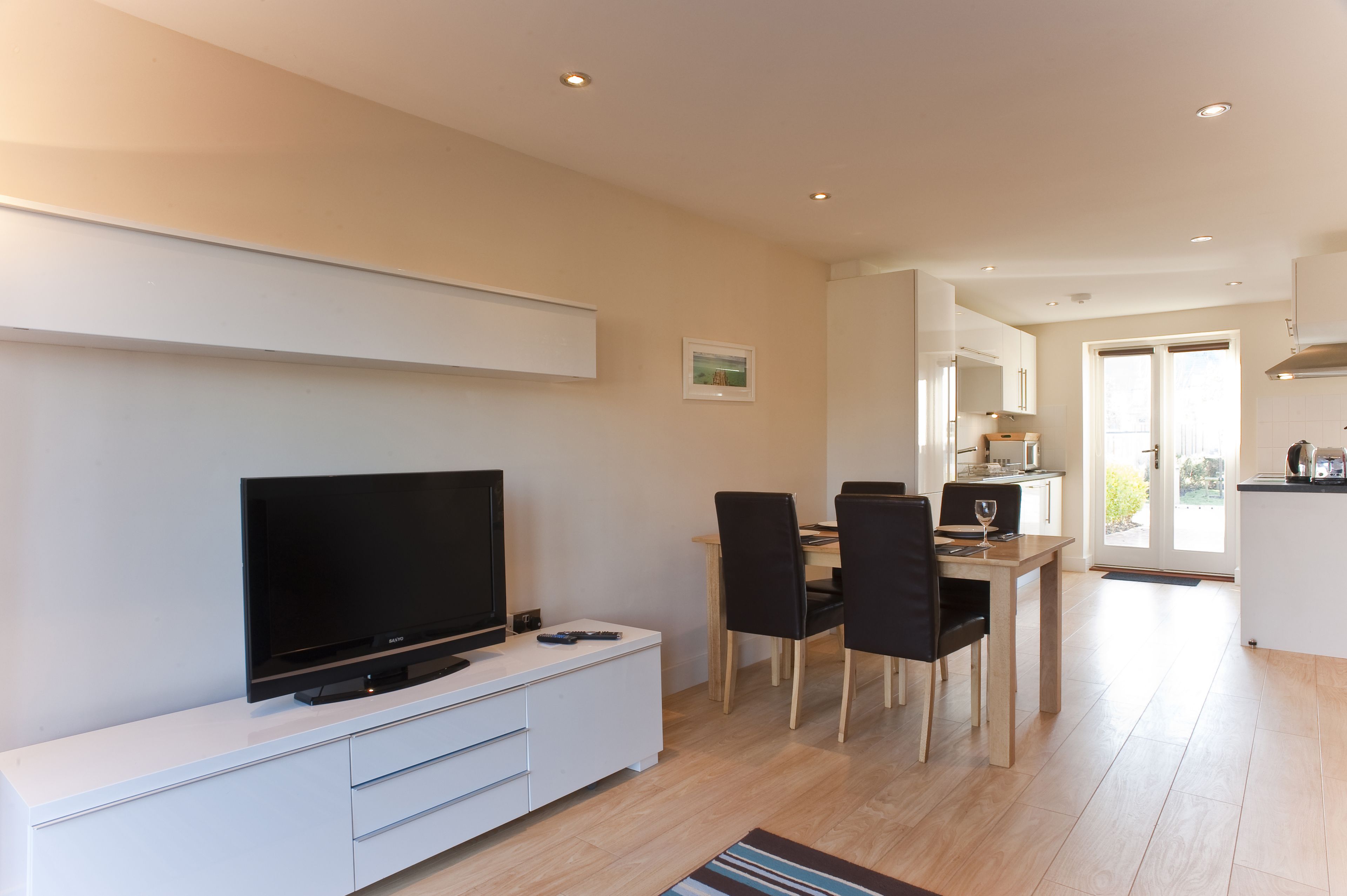 Un soggiorno moderno con una TV a schermo piatto su un mobile bianco, una zona pranzo con un tavolo in legno e sedie nere, e una cucina sullo sfondo.
