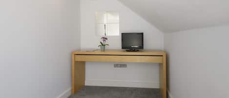 Una habitación pequeña y cerrada con techo inclinado, un escritorio de madera con televisor, una planta pequeña en maceta y un espejo en la pared.