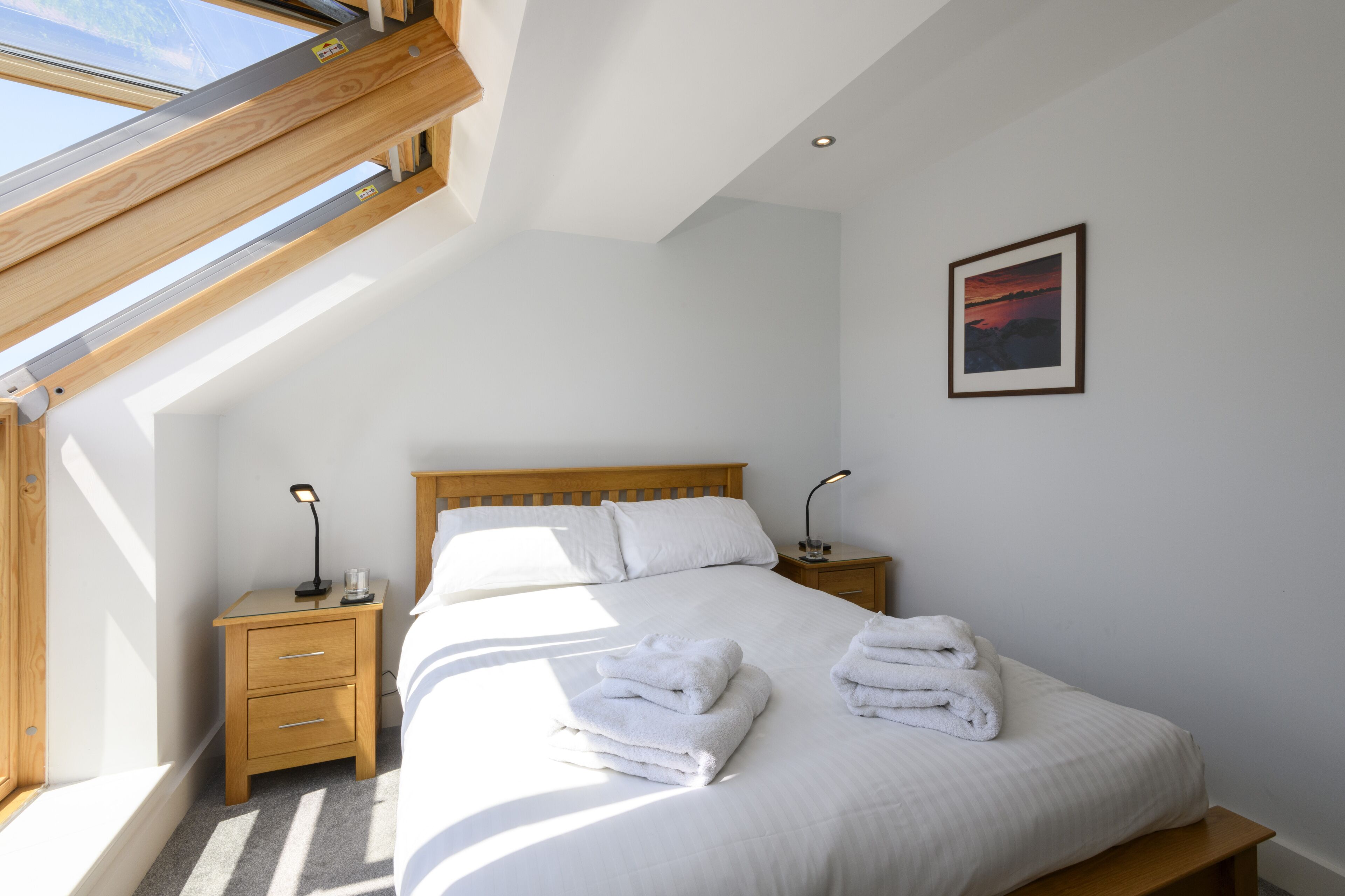 Un dormitorio con tragaluces, una cama con ropa blanca, dos mesitas de noche con lámparas y un cuadro en la pared.