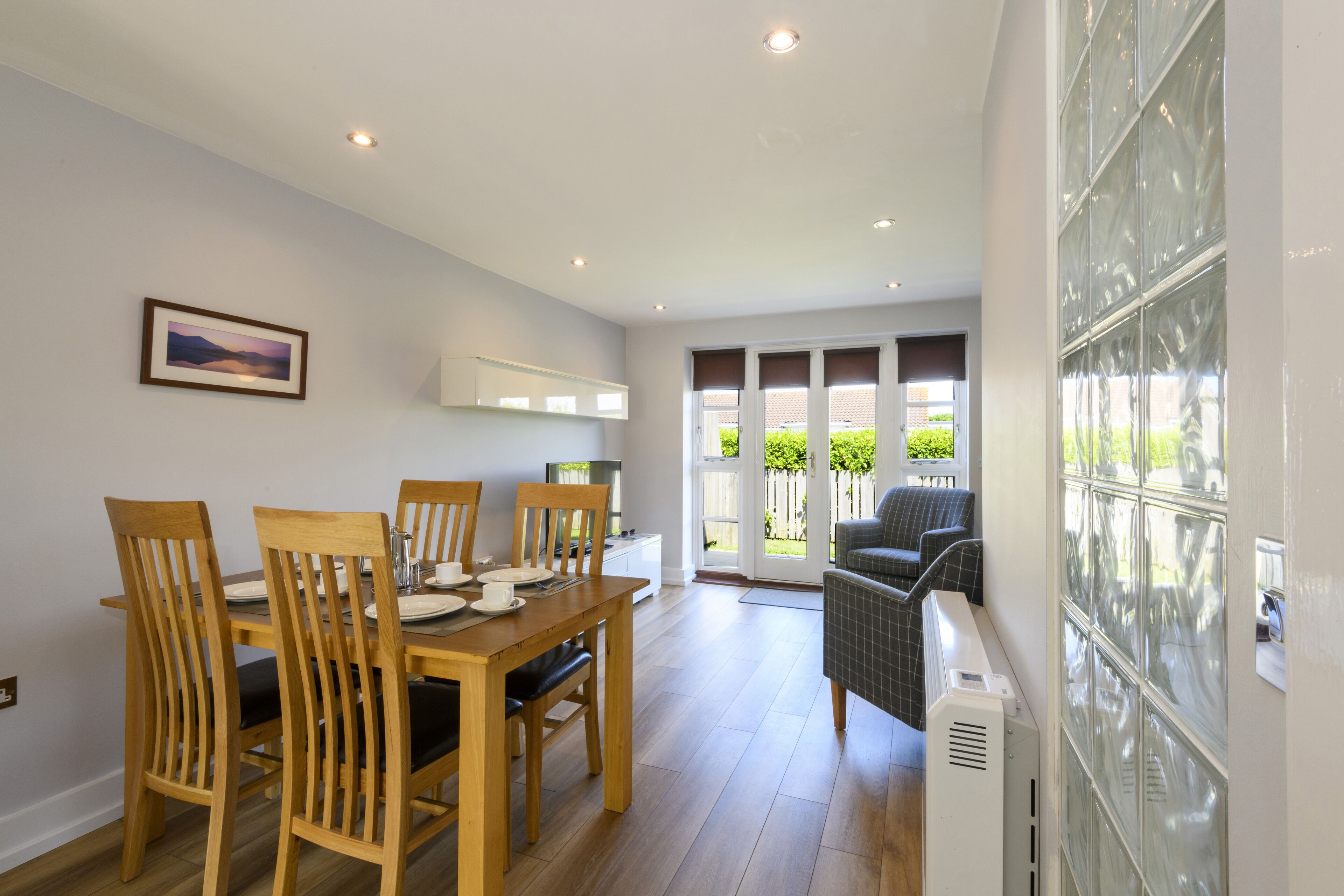 A dining area with a wooden table and chairs, a small kitchen area, and a view through glass doors to an outdoor space.