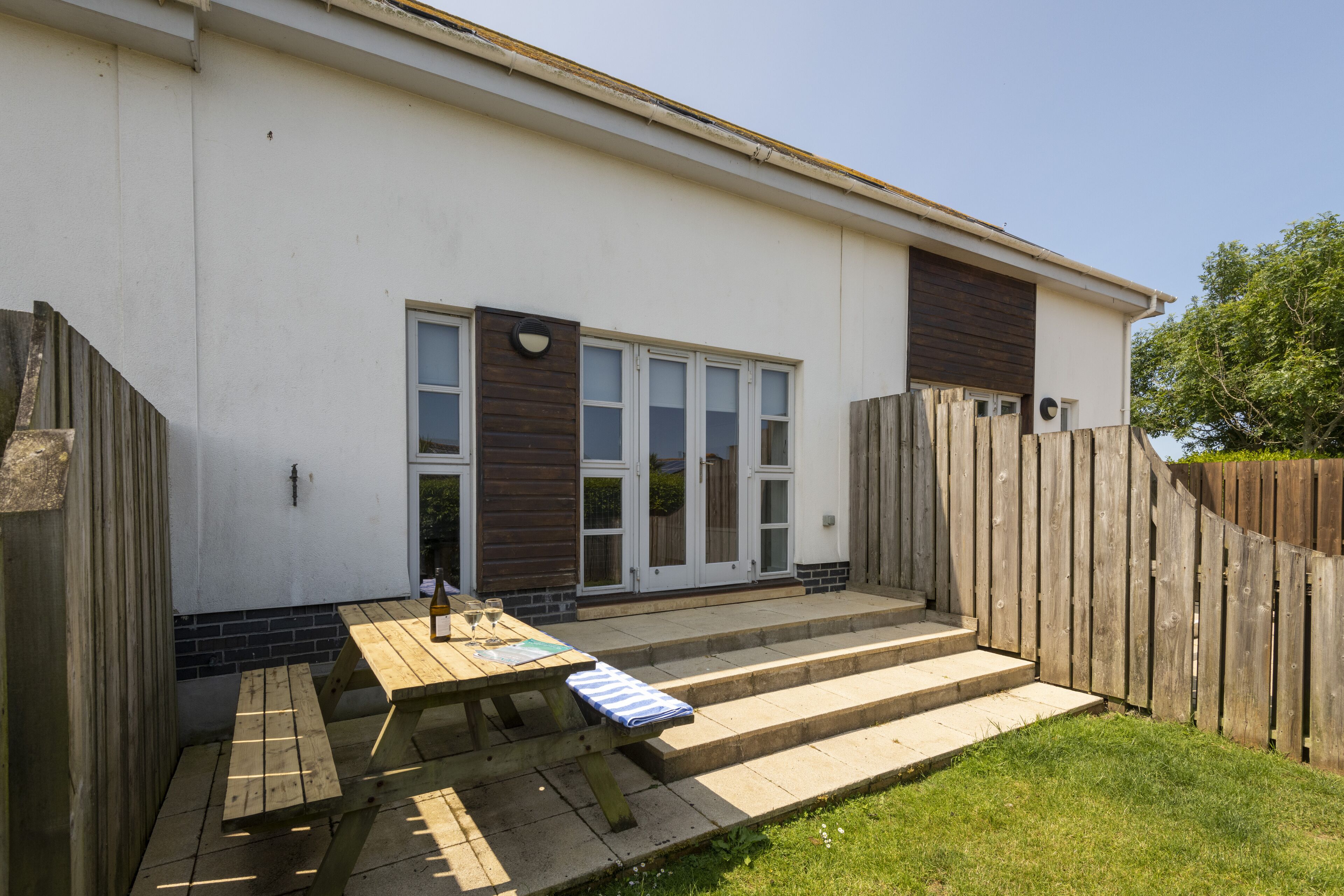 A patio with a wooden table and benches, a set of steps leading to a door, and a wooden fence surrounding the area.