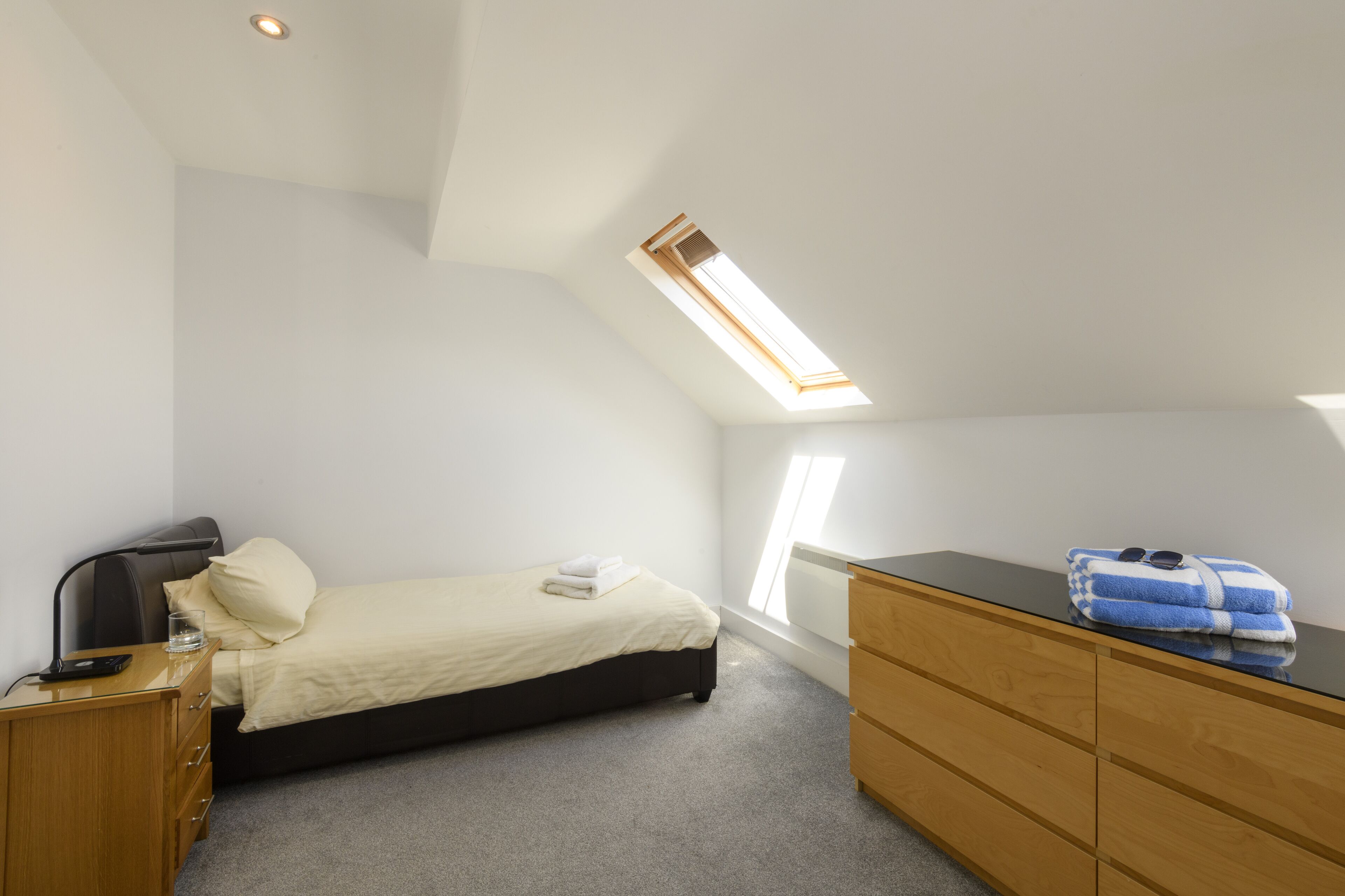Ein Schlafzimmer mit einem Bett, einer Holzkommode, einem Schreibtisch und einem Oberlicht.