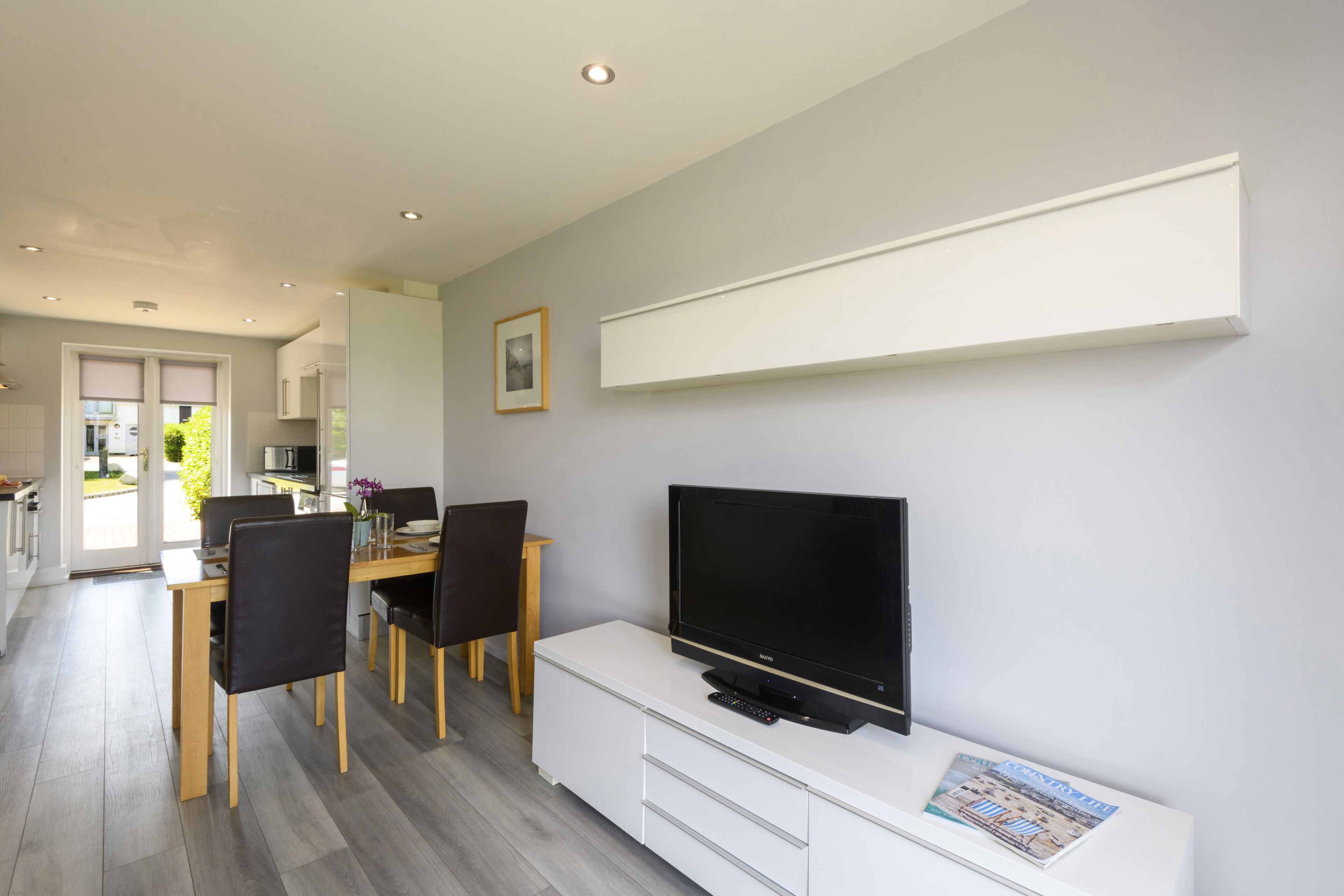 A modern living room with a flat-screen TV, a dining area with wooden chairs, and a kitchen visible through an open door.