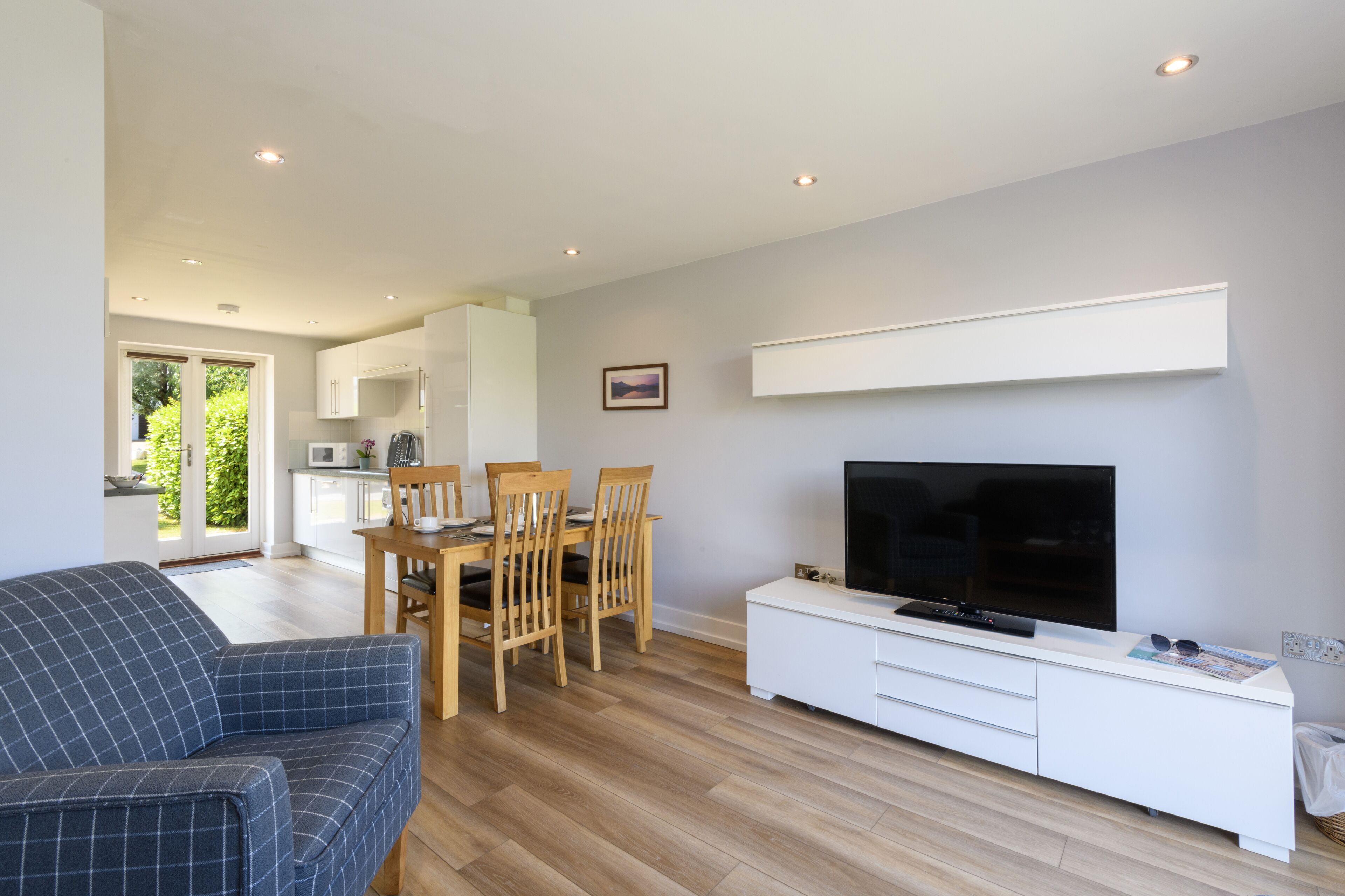 A modern living room with a sofa, a dining table with chairs, a flat-screen TV on a white cabinet, and a kitchen in the background.