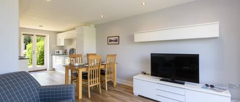 A modern living room with a sofa, a dining table with chairs, a flat-screen TV on a white cabinet, and a kitchen in the background.