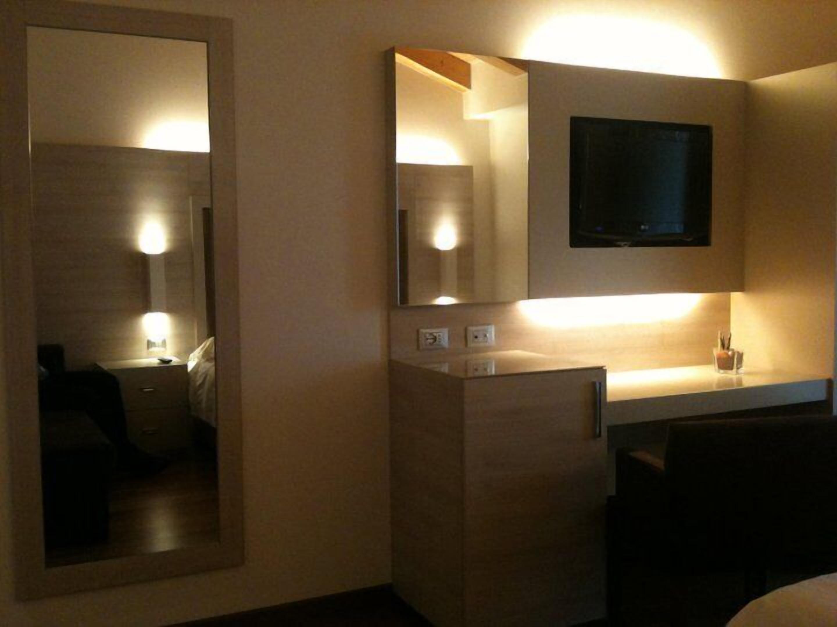 Quarto de hotel com um espelho grande, uma televisão de tela plana montada sobre uma cômoda de madeira e uma mesa de cabeceira com uma luminária.