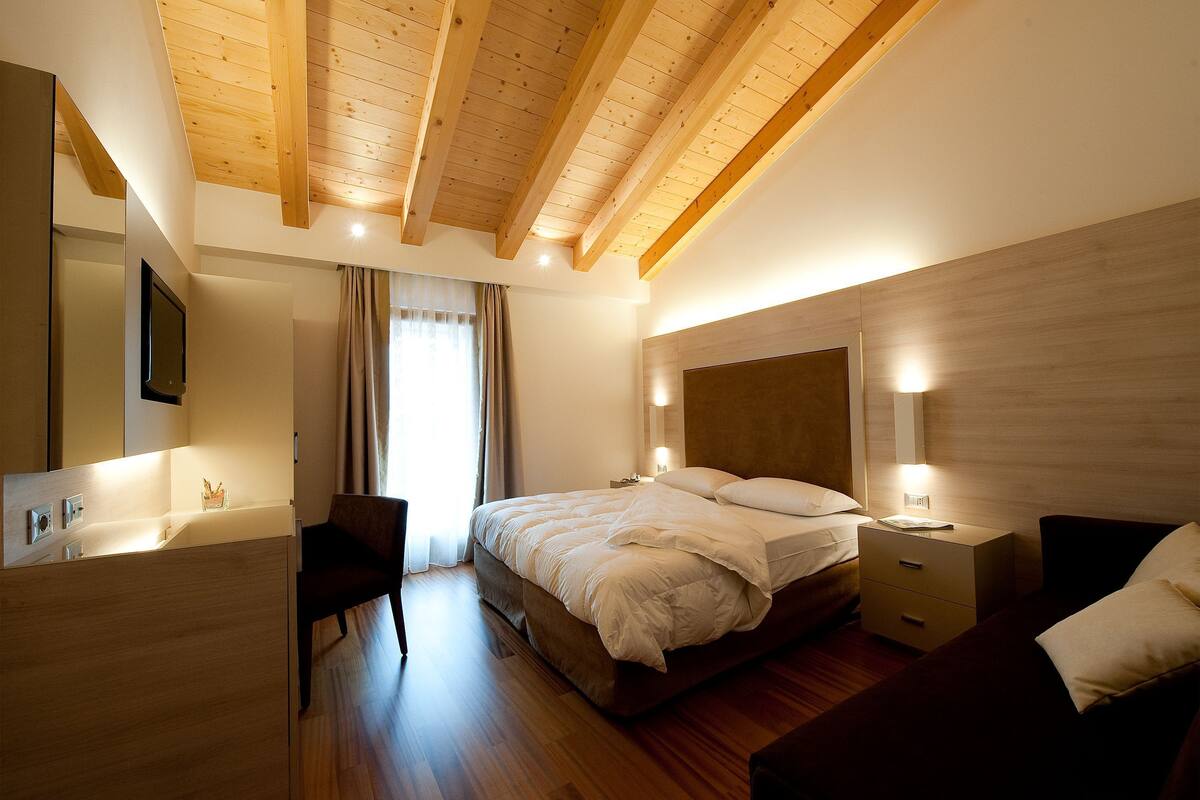 Un dormitorio con techo de madera, una cama con ropa blanca, una mesita de noche con lámpara y otro mueble auxiliar.