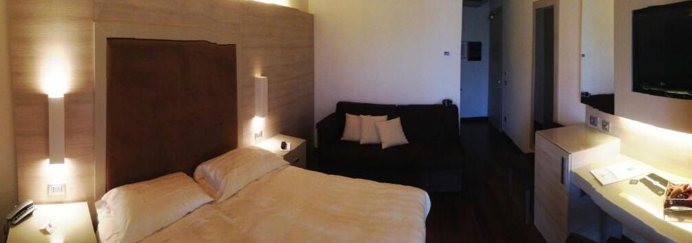 A hotel room with a bed, bedside table, lamp, and a small seating area with a couch and pillows.