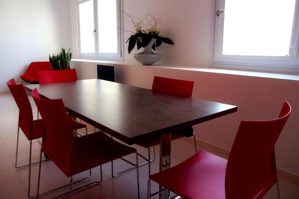 A dining area with a rectangular table and six red chairs. There is a potted plant on the windowsill.