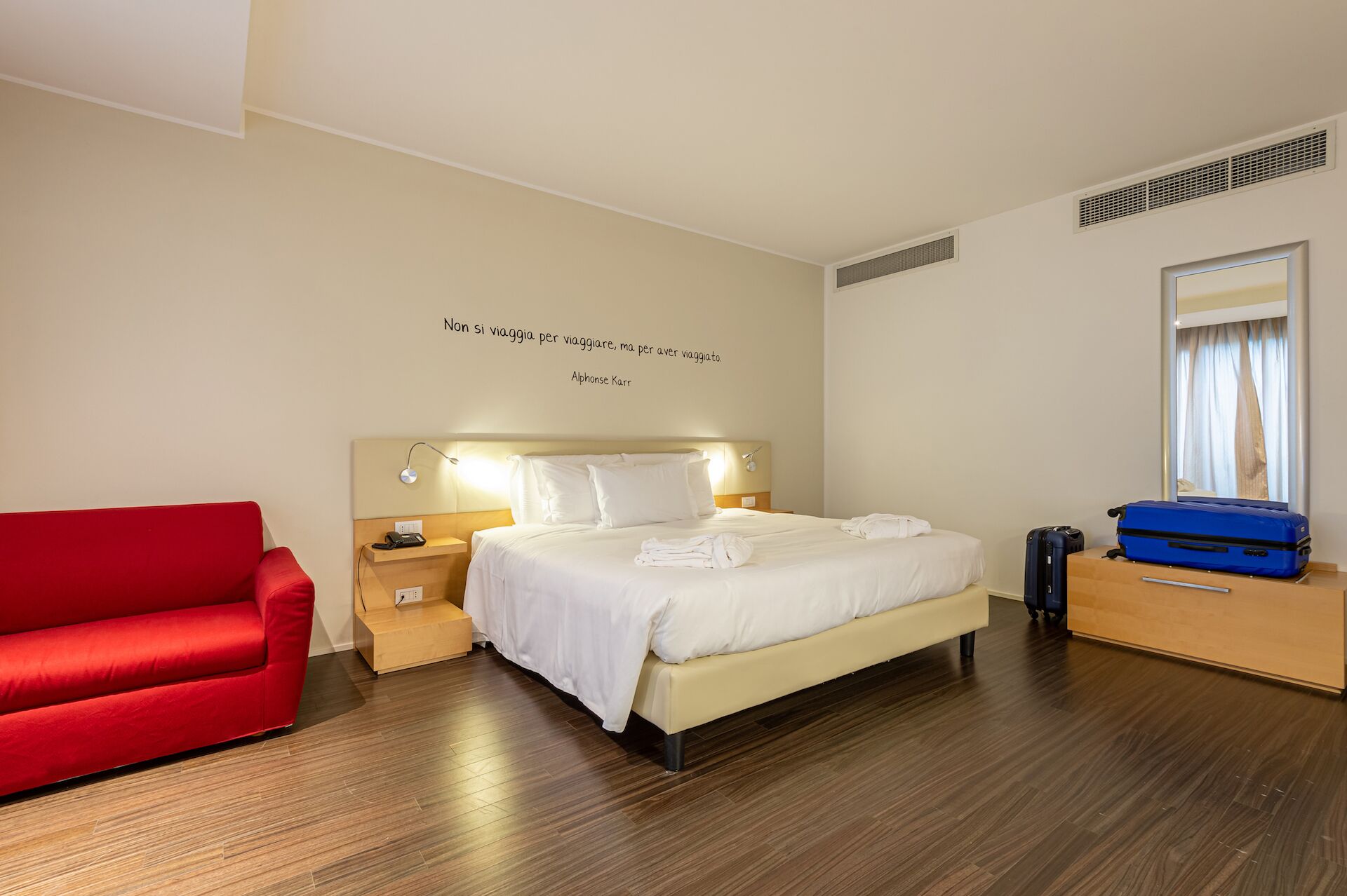 A hotel room with a bed, a red armchair, a wooden dresser, and a blue suitcase.