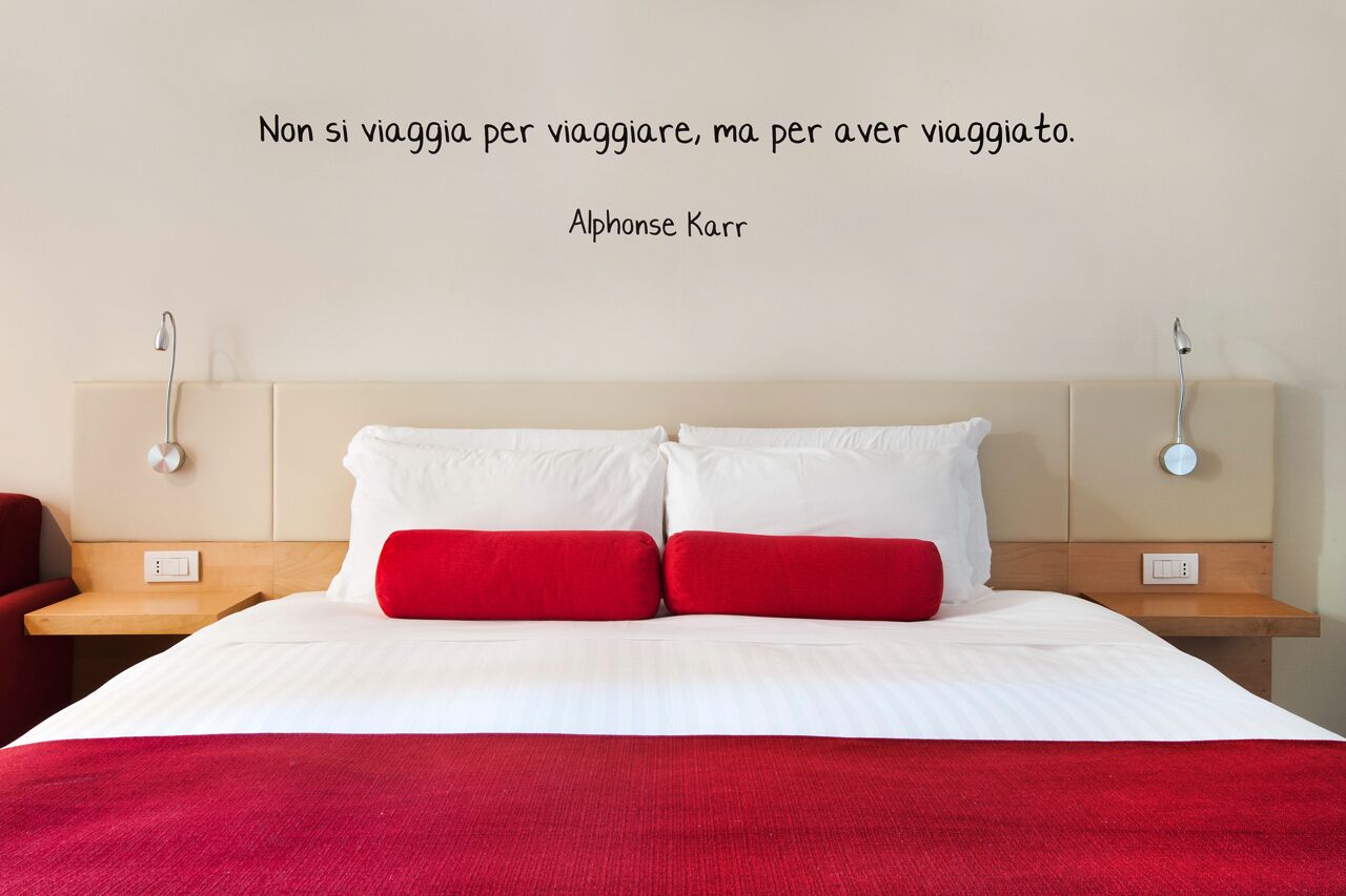A neatly made bed with white and red bedding, a wall quote by Alphonse Karr, and bedside lamps.